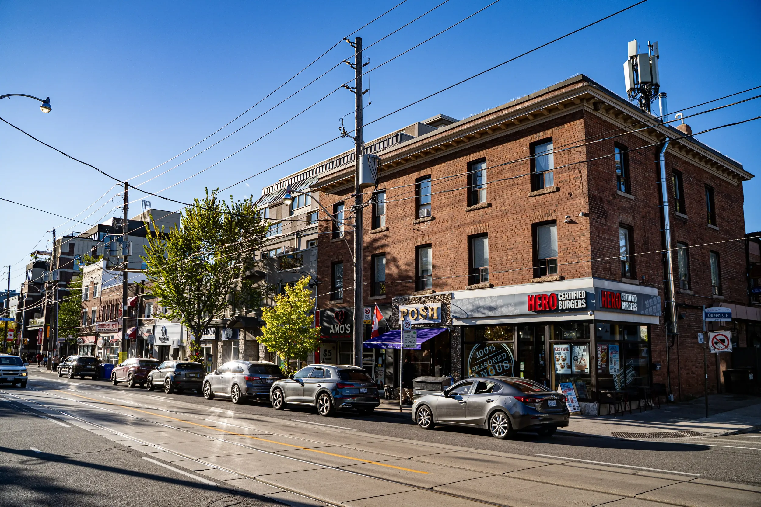 City street scene with parked cars, brick buildings, storefronts, and overhead power lines against a clear blue sky.