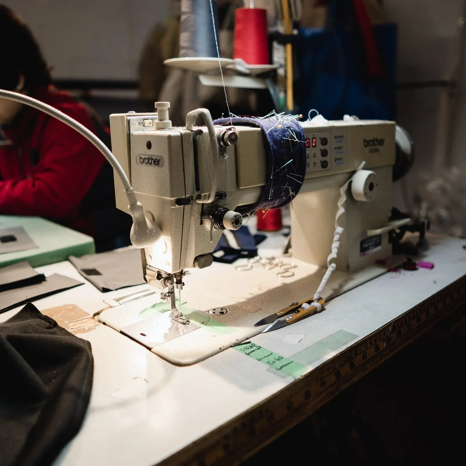 A sewing machine on a worktable surrounded by fabric, scissors, and fabric pieces. The machine is actively sewing a piece of dark blue fabric with pins attached.