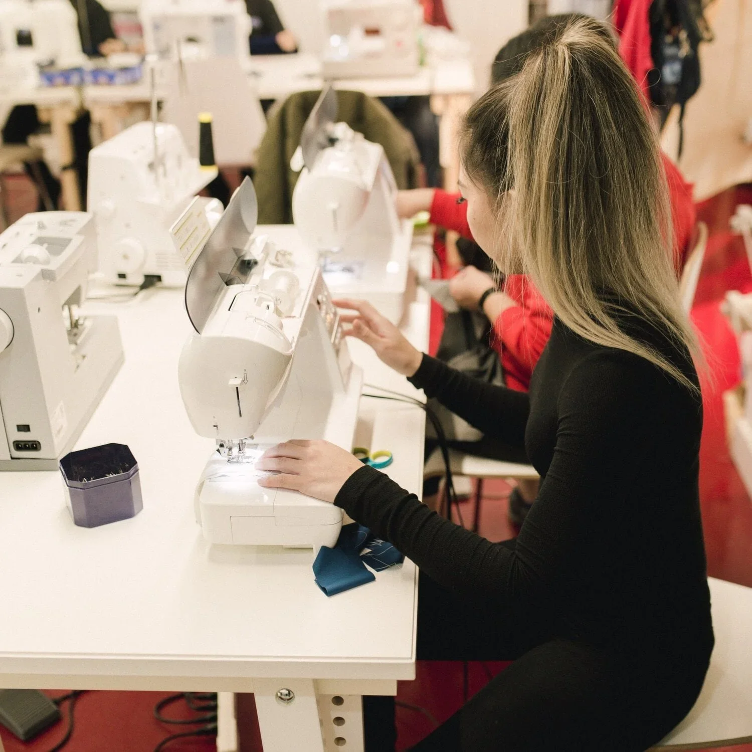A woman sewing on a white sewing machine at a white table with other sewing machines and materials, in a sewing or craft workshop.