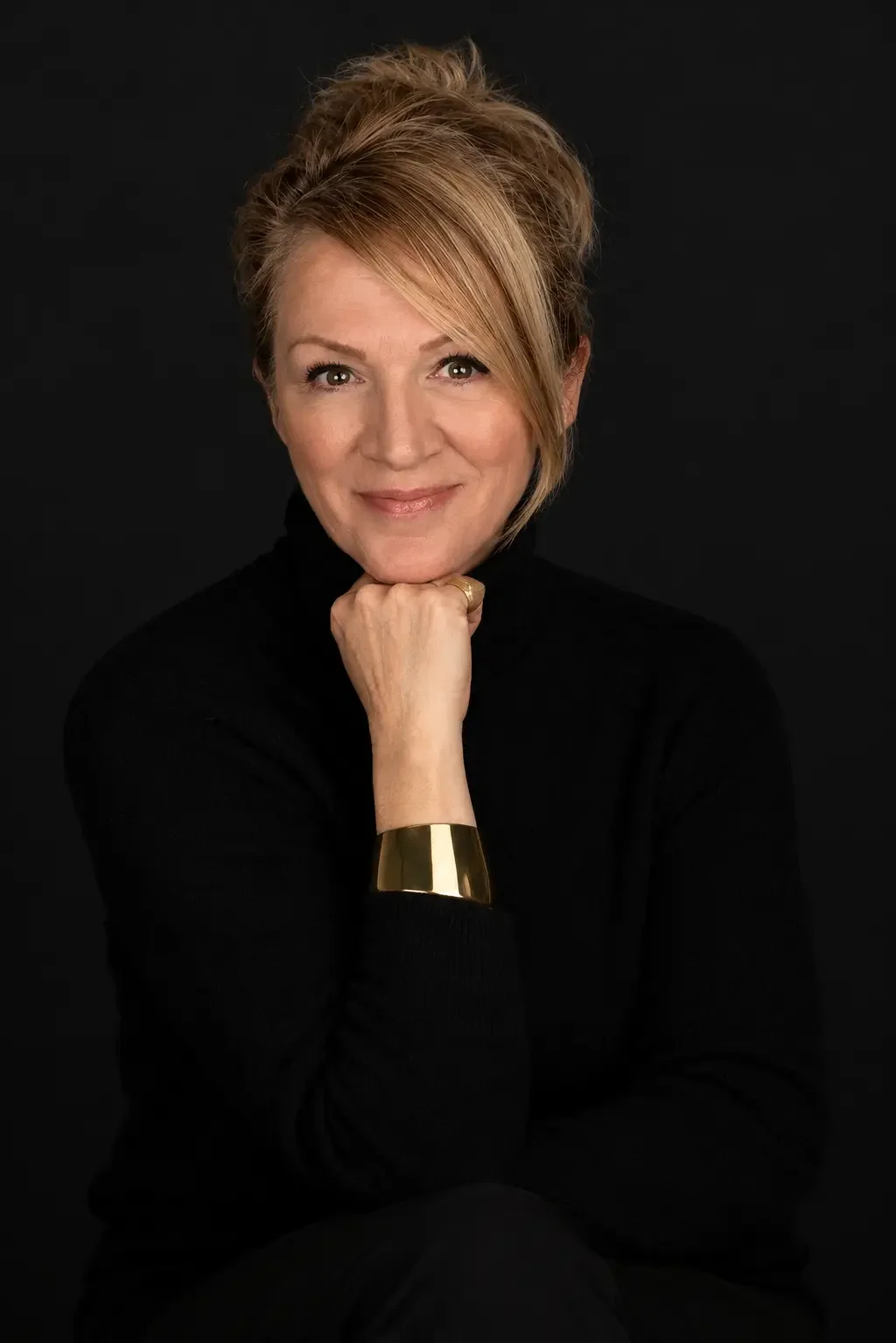 Portrait of a woman with short blonde hair, smiling, wearing a black turtleneck and gold bracelet, posed against a dark background.