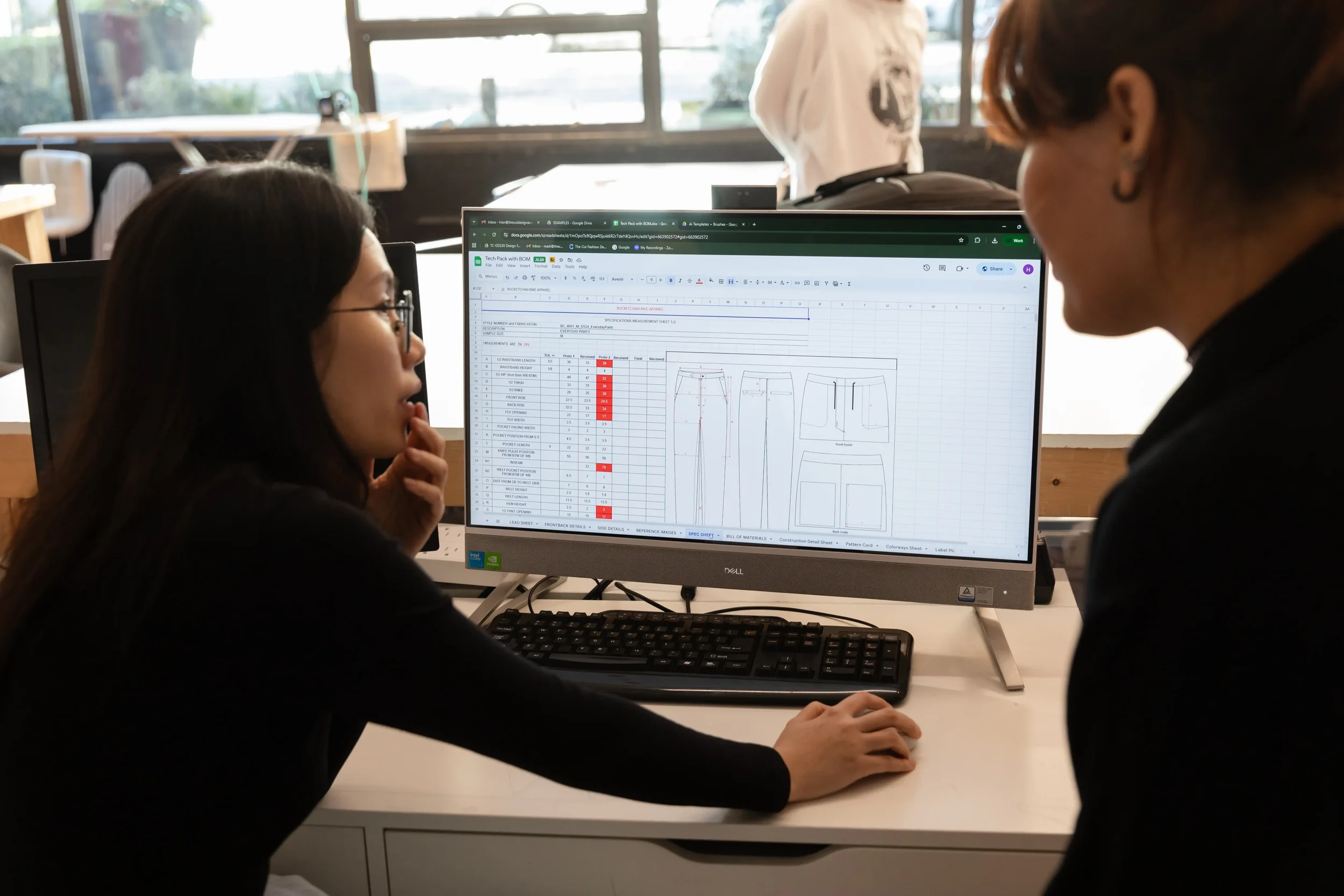 Two women looking at a computer screen displaying technical data and sketches, in a bright office with large windows.