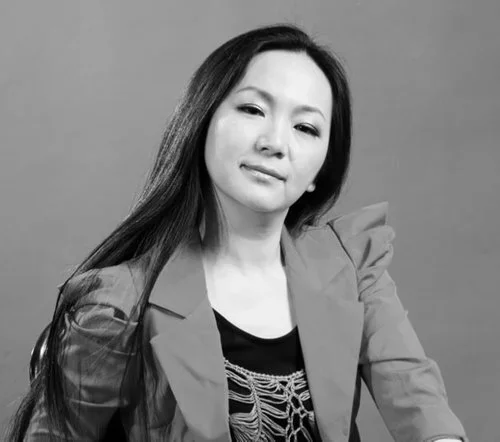 A woman with long dark hair wearing a blazer and a graphic t-shirt, posing with a confident expression against a plain background.