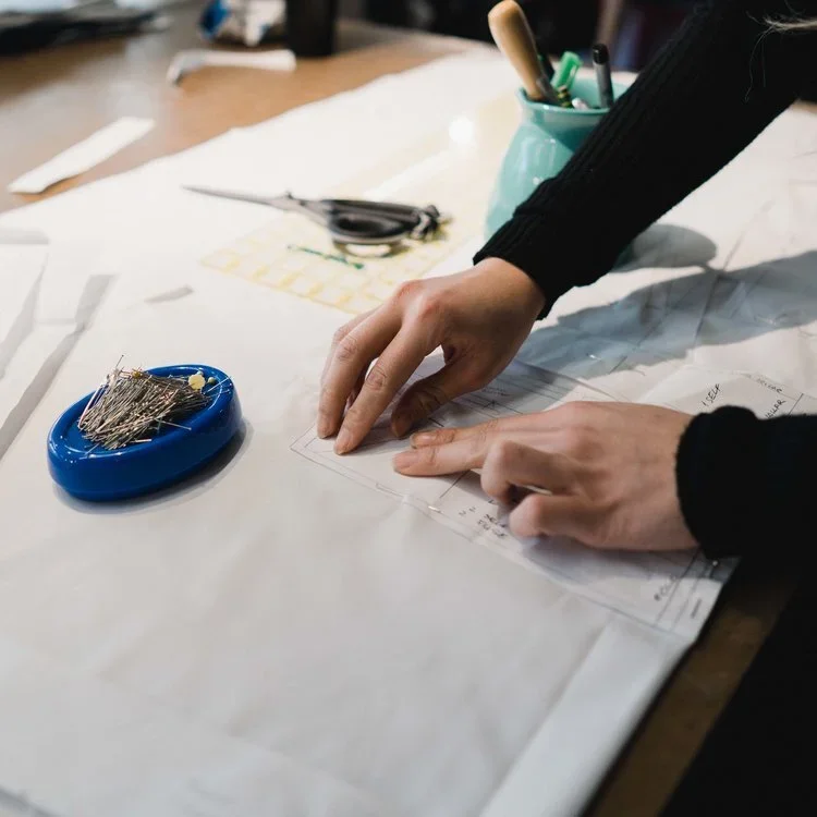 Person working on a sewing project, pinning pattern paper on a table with sewing pins in a blue container and scissors in the background.