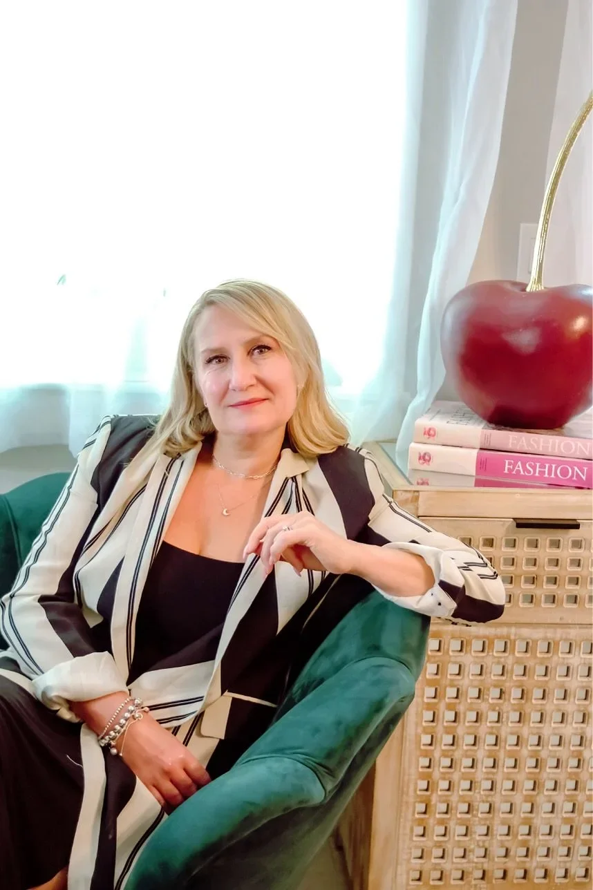 A woman with blonde hair sits on a green velvet chair in a well-lit room. She is wearing a black top and a black-and-white striped blazer. Behind her, there is a window with sheer white curtains. To her right, a side table holds a large red cherry-shaped decorative object and three pink fashion books.