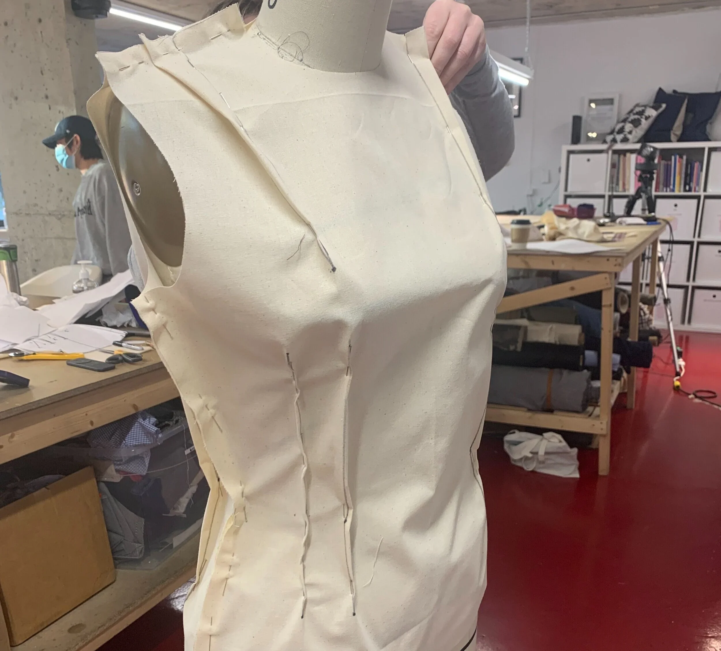A dress form on a work table in a workshop, with fabrics, scissors, and tools around. Another person wearing a mask and cap is in the background, working or observing.