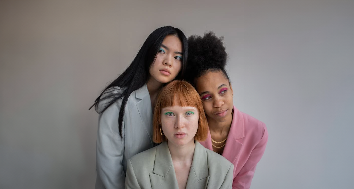 Three women with colorful makeup and stylish blazers posing together against a plain background.