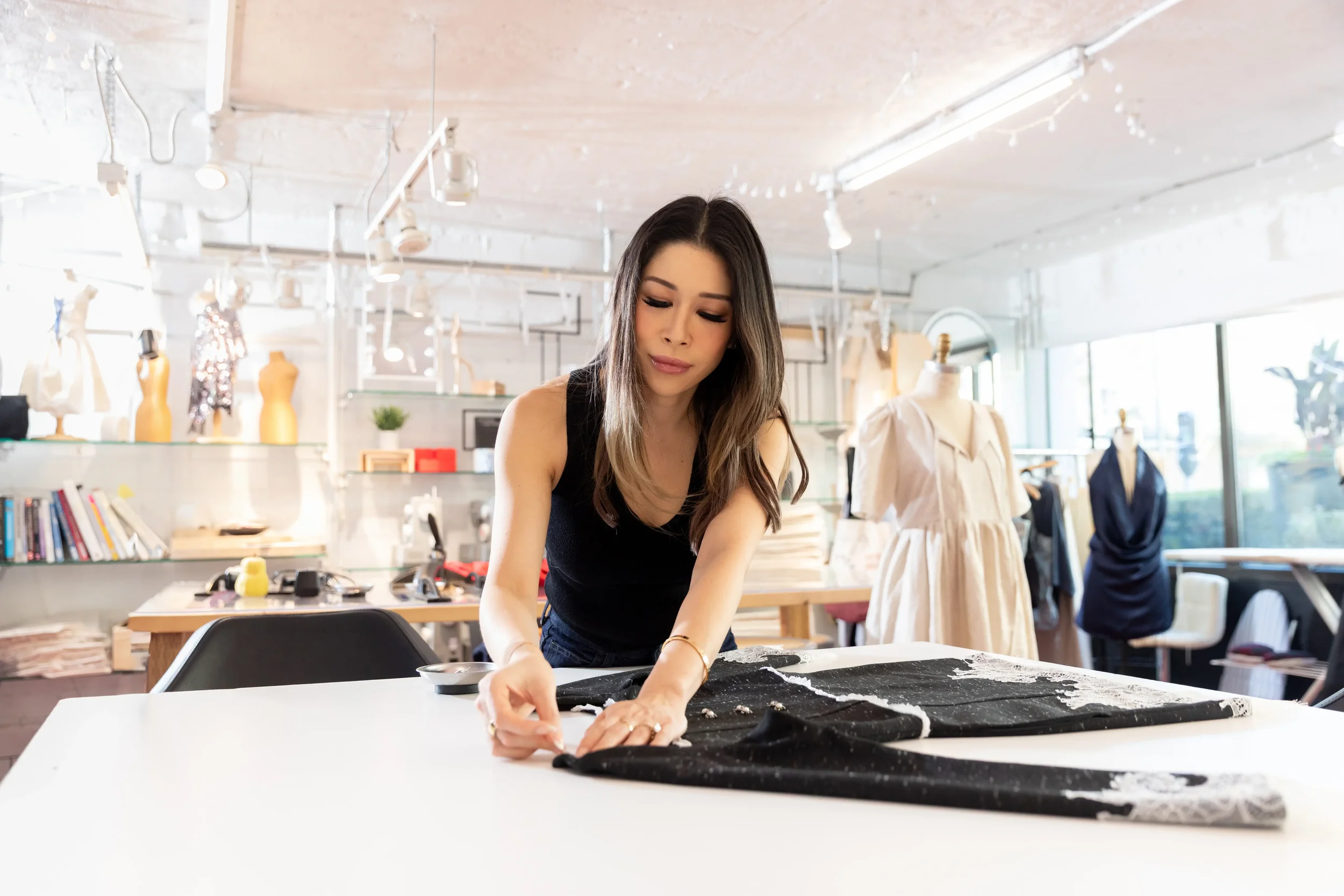 A woman working on a fabric project at a workshop table in a bright, modern space filled with mannequins and sewing supplies.