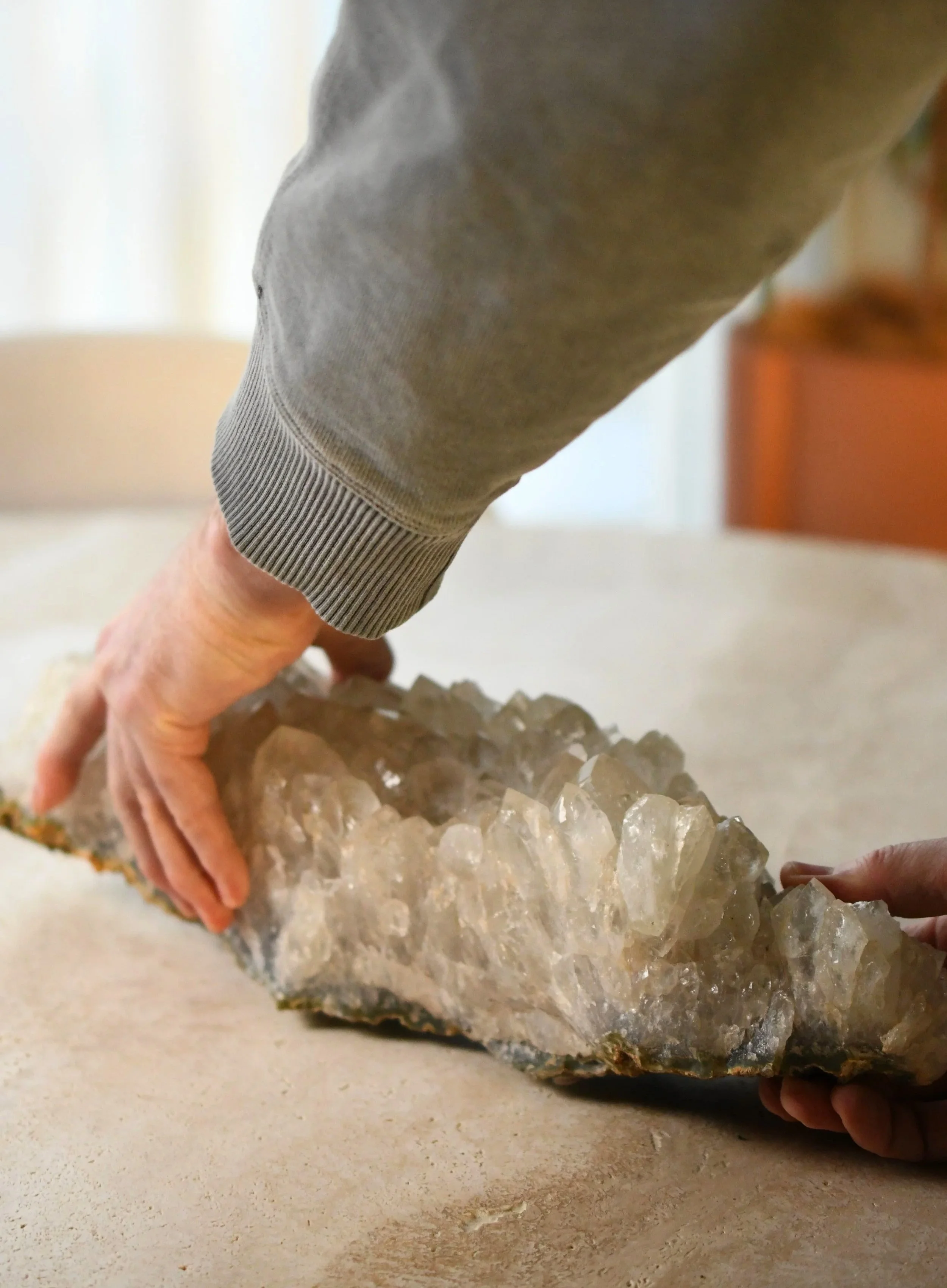 Close-up of a person with gray sleeves touching a large, rough crystal on a beige surface.