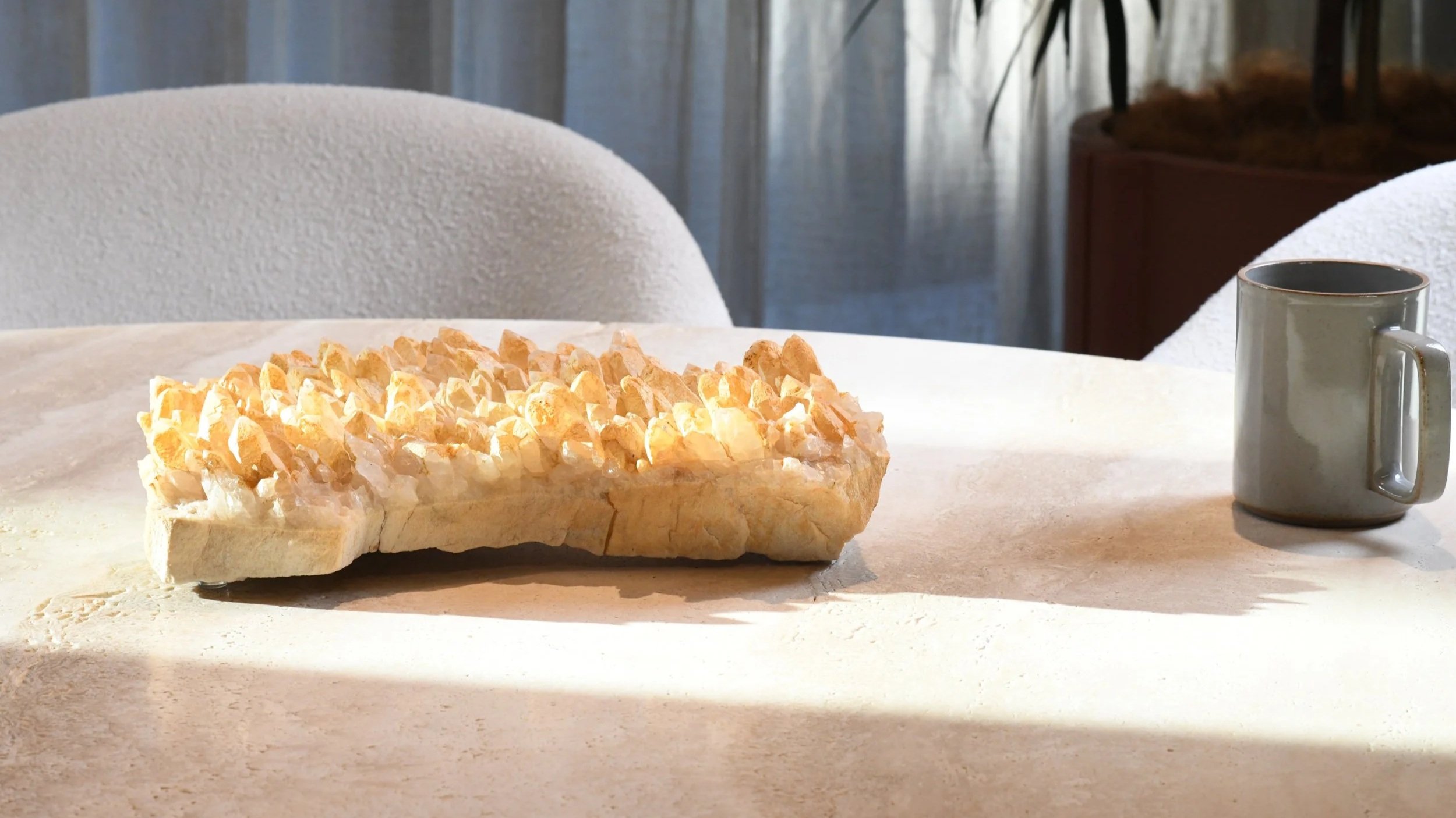 A crystal with a textured surface is placed on a white countertop, alongside a grey ceramic mug and a blurry background with plants and curtains.