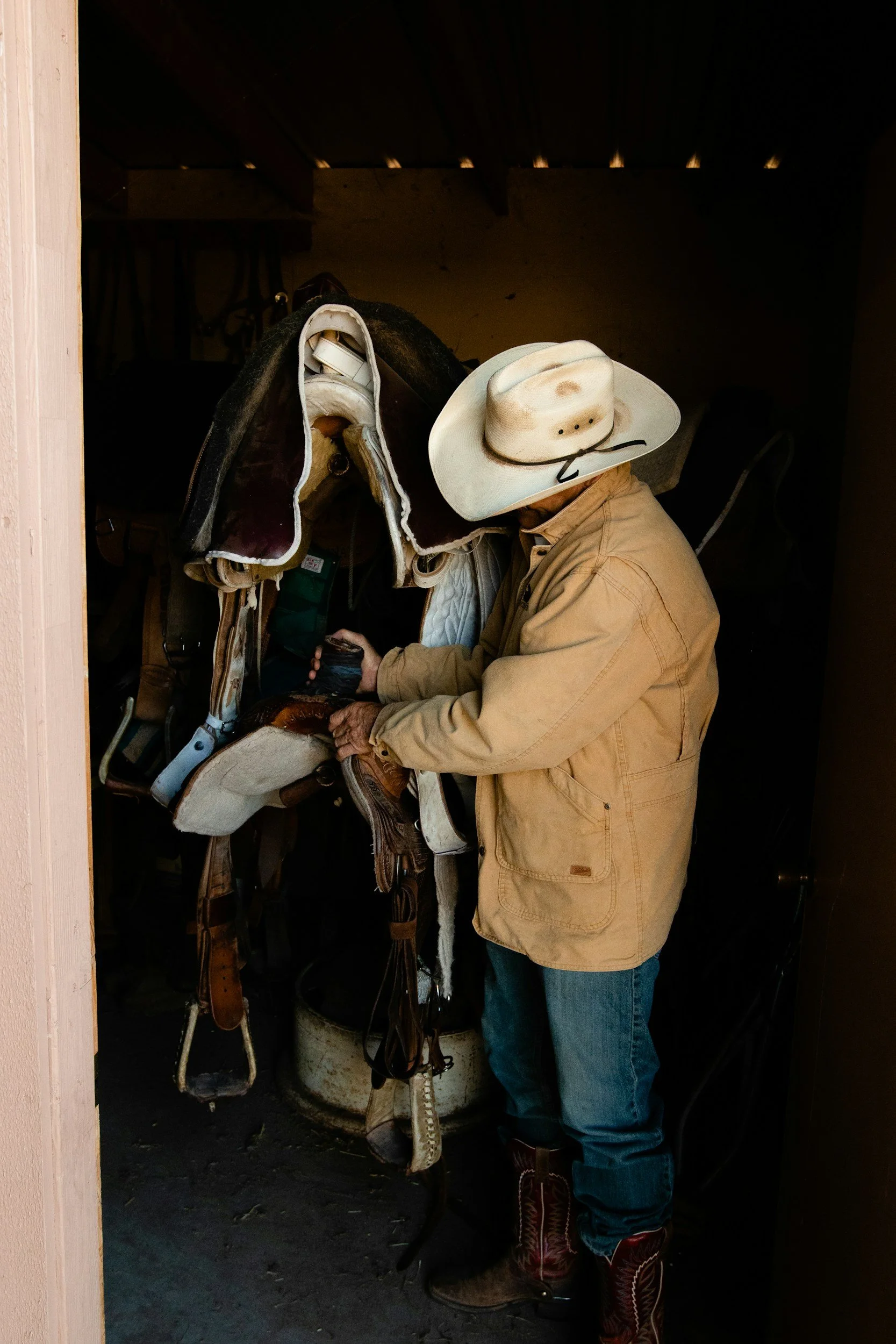 A person wearing a cowboy hat and cowboy boots is examining a horse saddle in a dark room.
