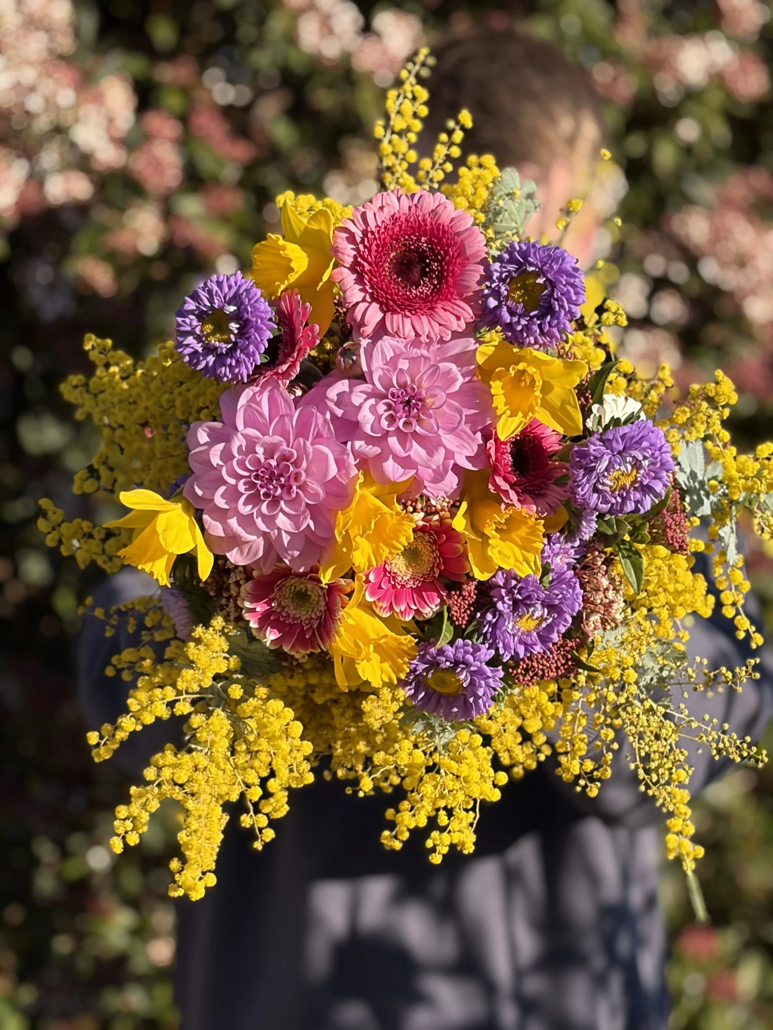 A person holding a colorful bouquet of flowers including pink, yellow, purple, and white blossoms, with a blurred background.