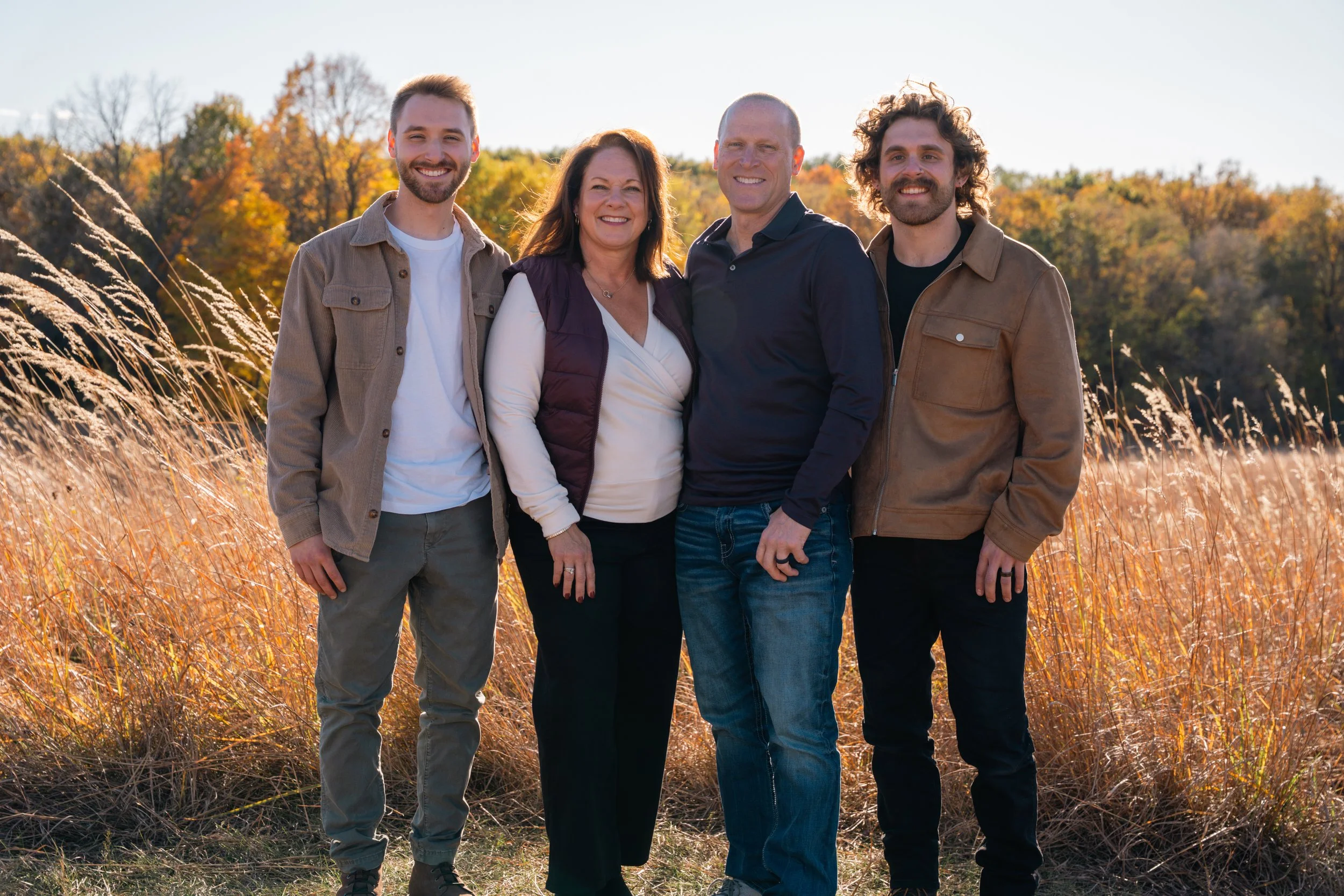 Group of four people standing outside in a field of tall grass with autumn-colored trees in the background, smiling at the camera.