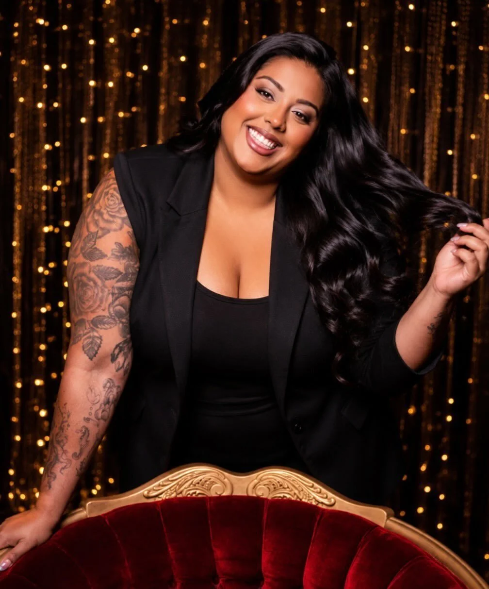 A woman with long dark hair wearing a black outfit and blazer, smiling and holding her hair, standing behind a red velvet chair, with a backdrop of gold sparkling curtains.