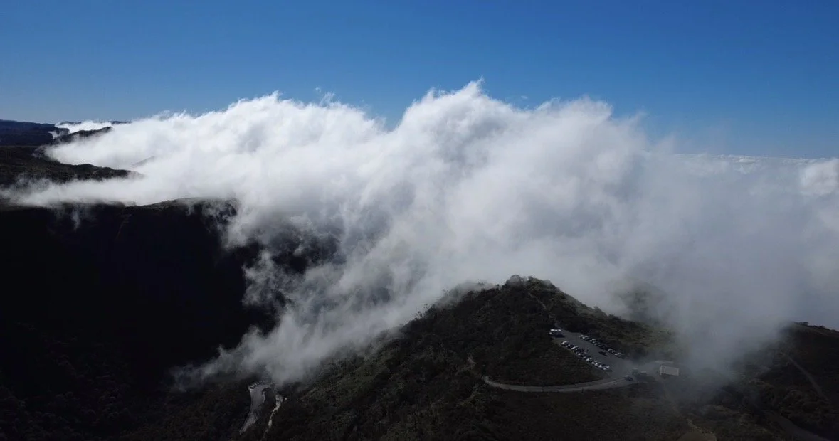 Clouds covering a mountain landscape with a parking lot, under a clear blue sky.