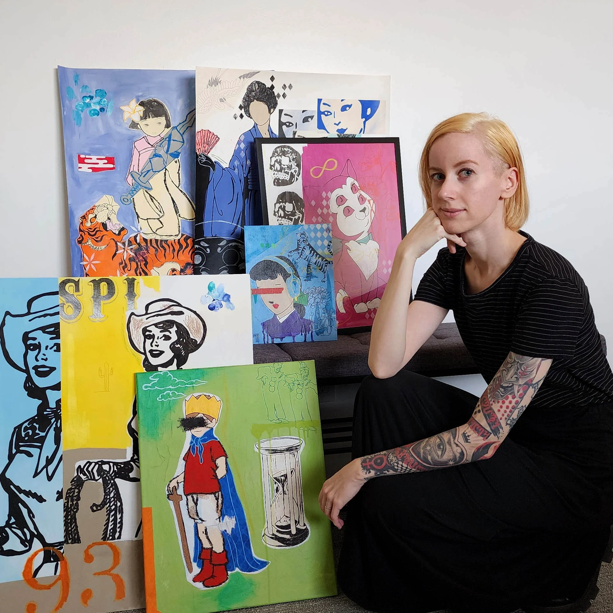 Young woman with short blonde hair and tattoos on her arm, sitting beside a collection of vibrant artwork featuring portraits of women and artistic designs on a white wall.