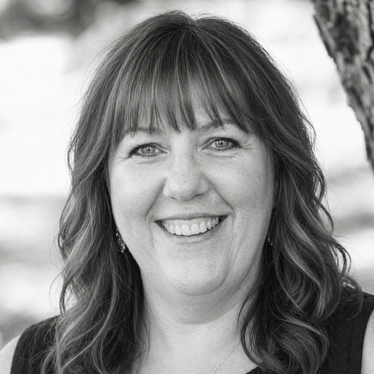 Close-up black-and-white portrait of a woman with wavy hair, smiling, outdoors with a tree in the background.