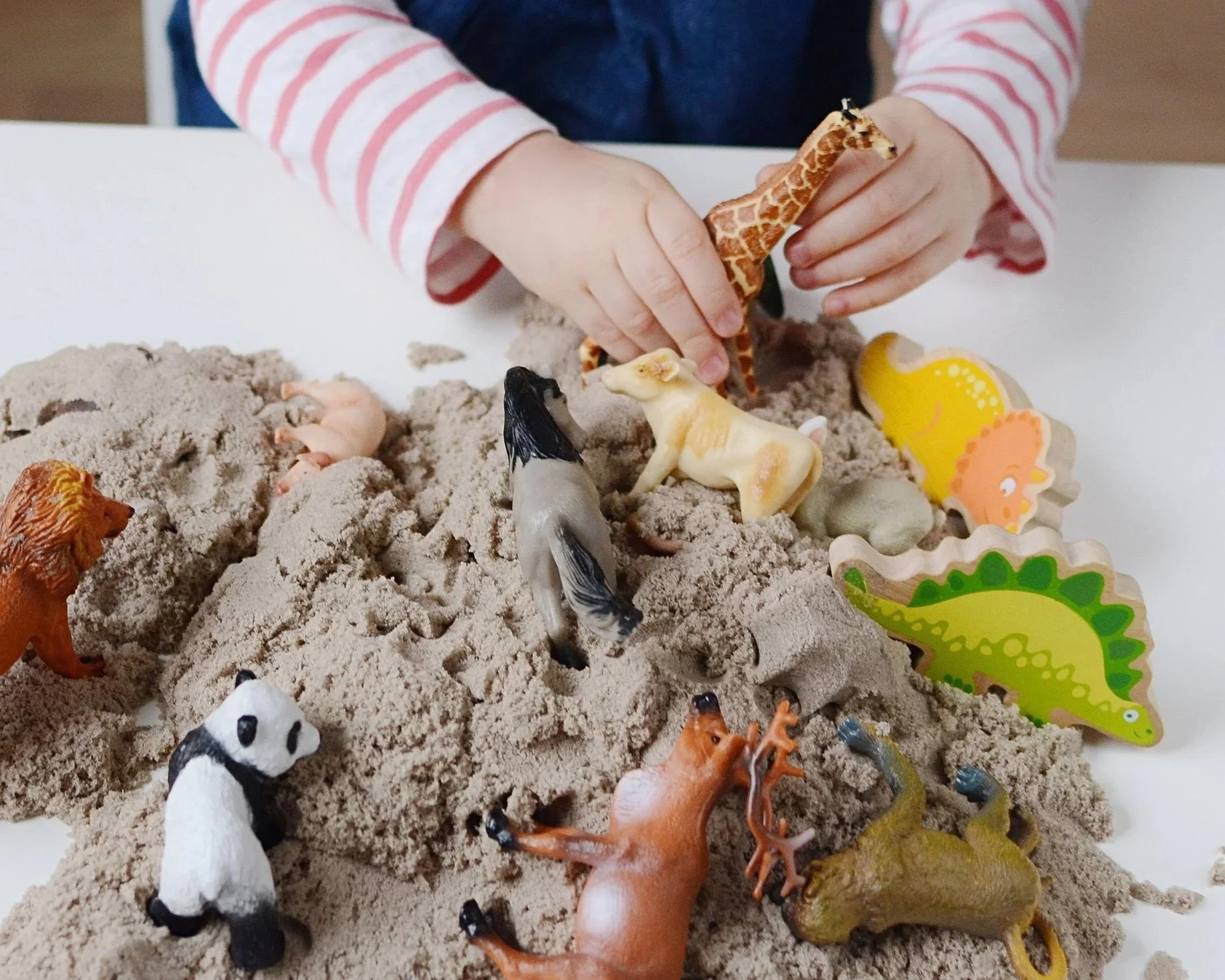 Child playing with toy animals in a sandbox, including giraffes, zebras, lions, and other animals, with colorful animal-shaped toys around.