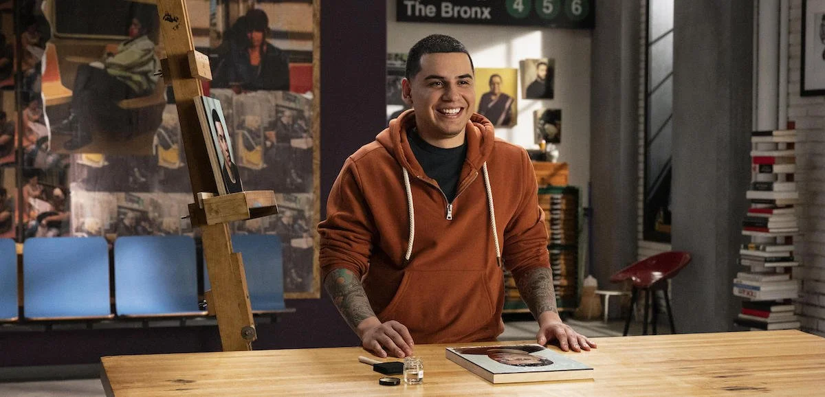 A smiling young man with tattoos wearing a brown hoodie standing behind a book on a wooden table in an indoor space with bookshelves and artwork in the background.