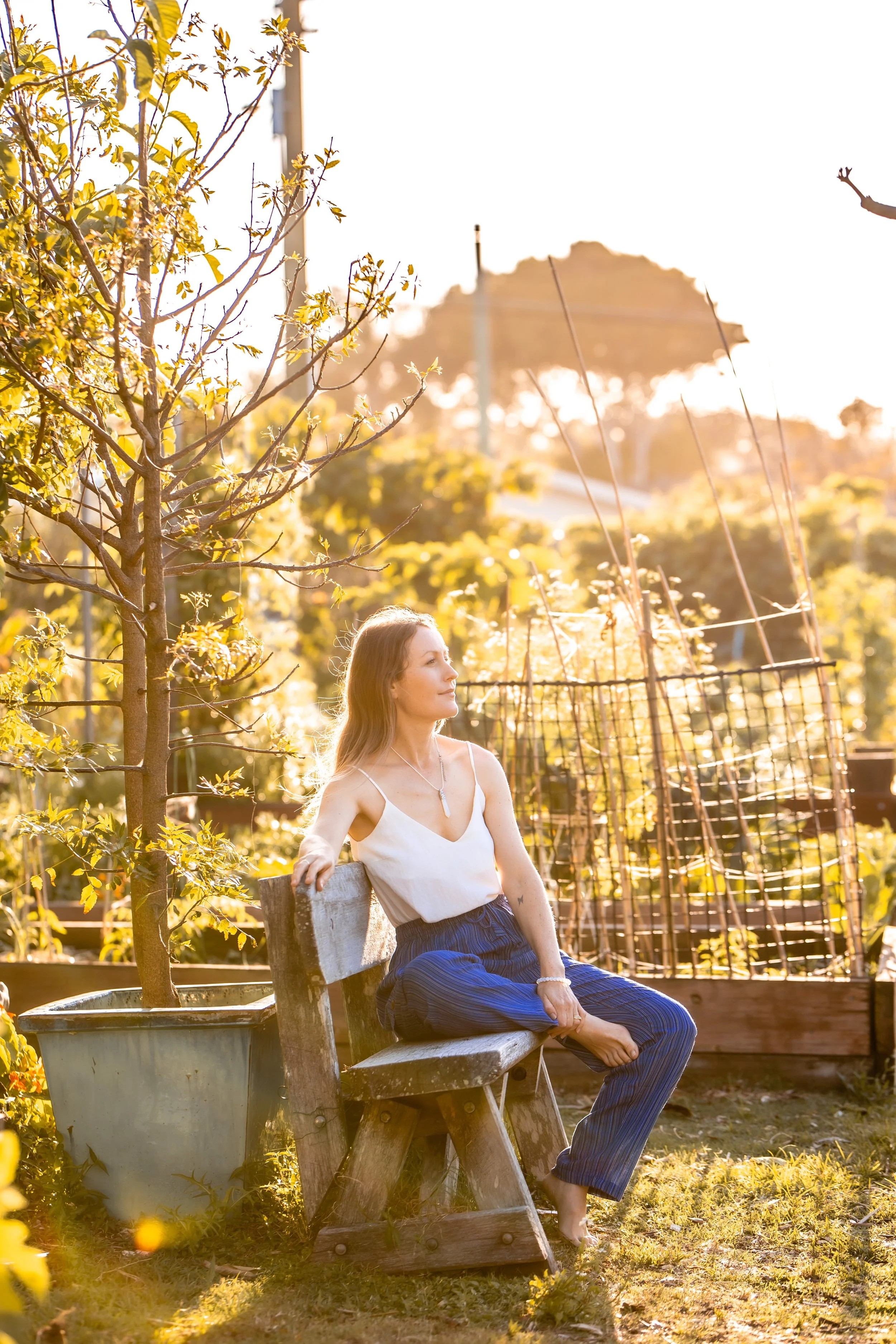 A woman with long hair and tattoos sitting on a wooden garden bench, wearing a white top and blue pants, surrounded by trees and plants in a garden during golden hour sunlight.