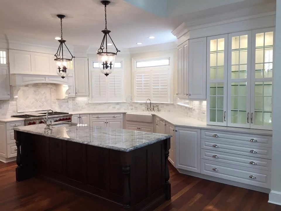 Elegant white kitchen with marble countertops, dark wood island, glass-front cabinets, pendant lights, and stainless steel range.