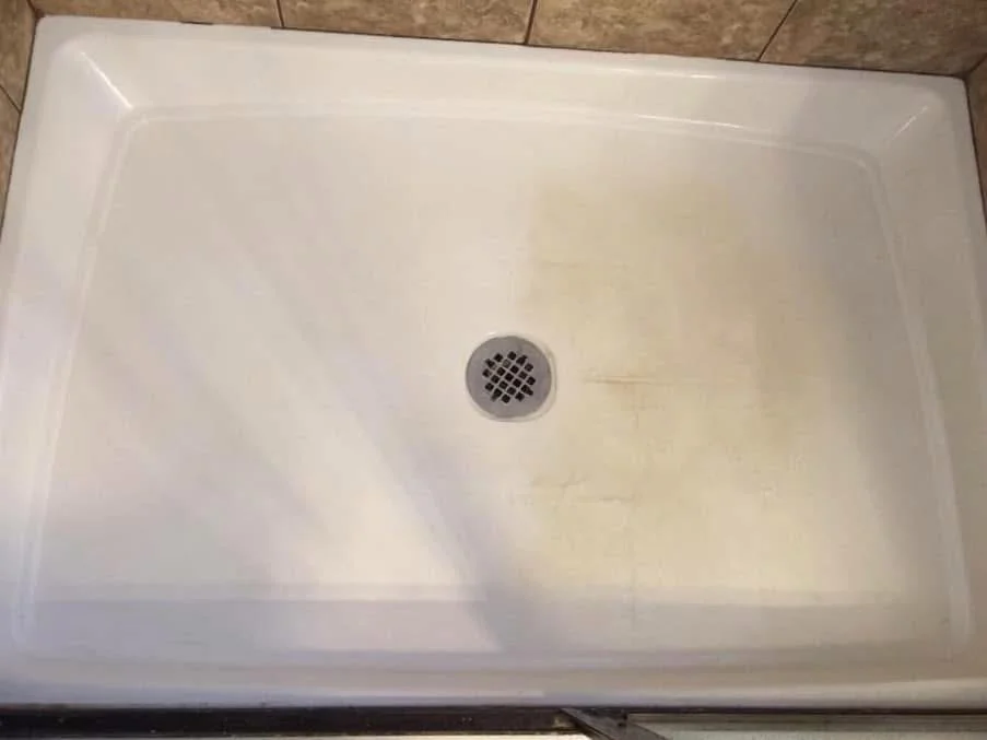 White shower pan with a metal drain and visible yellowish stains against a tiled wall