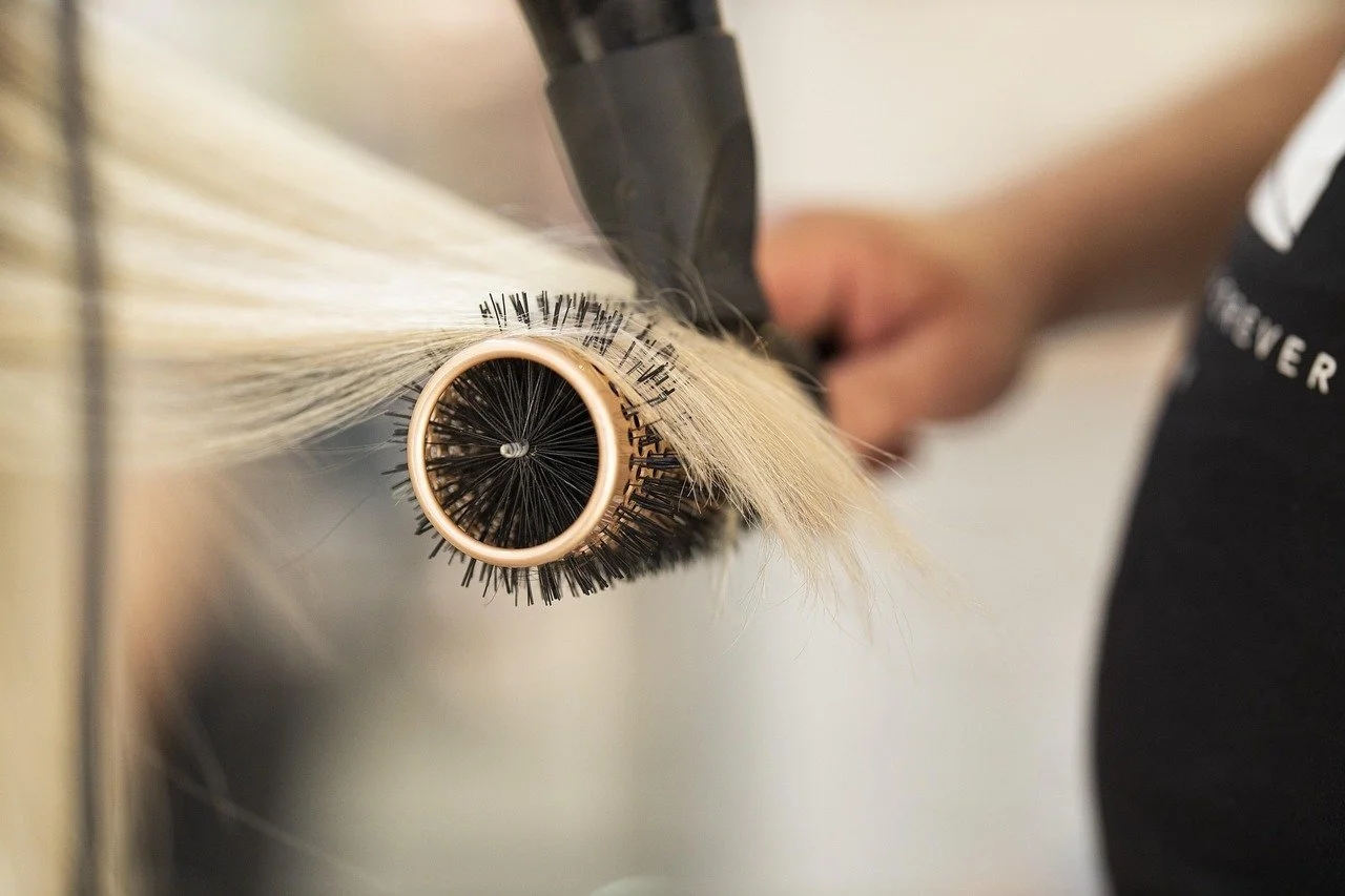 A person applying eyelash extensions using a curling tool. The focus is on the eyelash extension process, with the person's hand holding the tool close to a roller holding blond synthetic eyelashes.