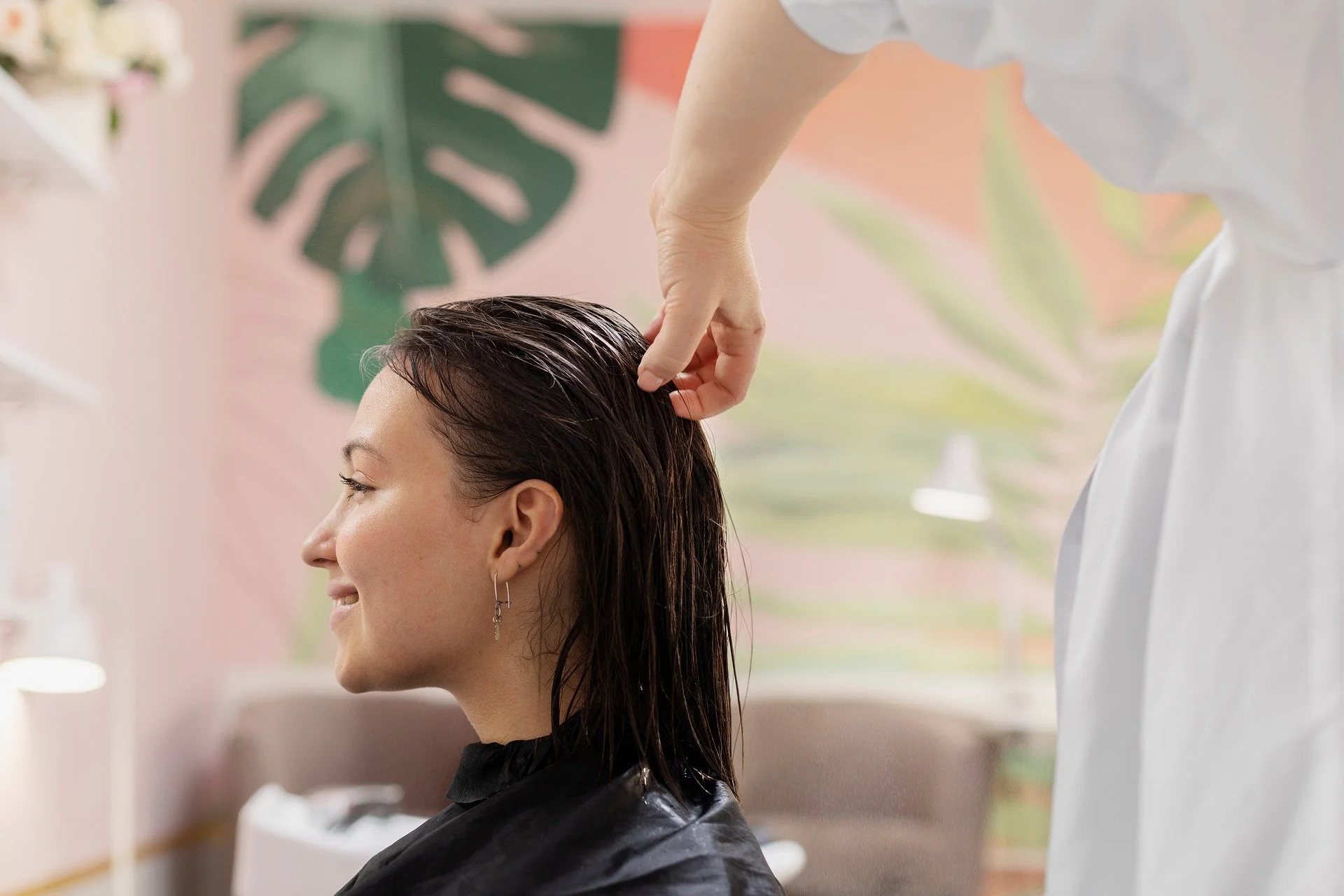 A woman with dark hair getting her hair styled or treated by a professional in a salon, with a pink and green painted wall in the background.
