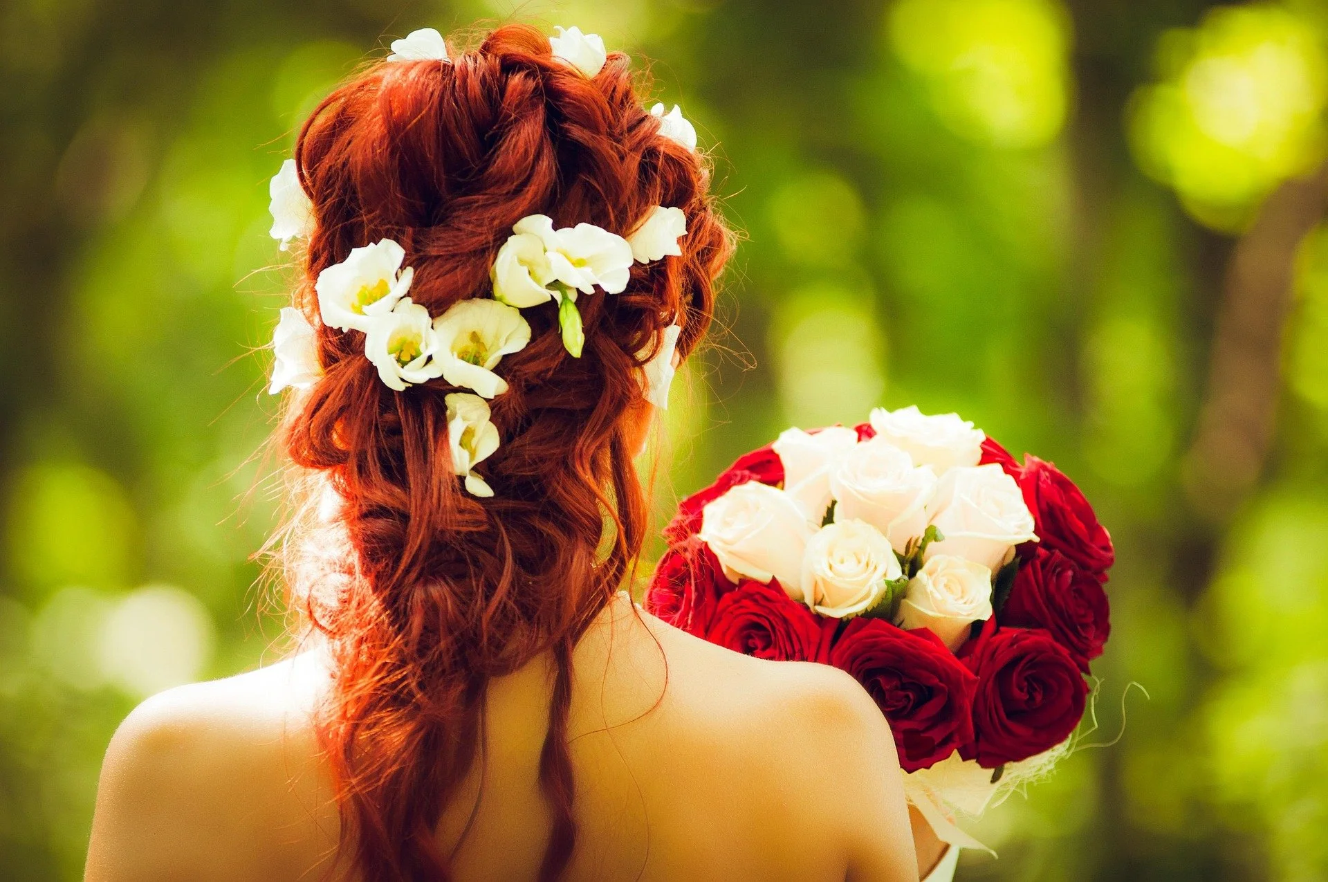 Back of a woman with long red hair decorated with white flowers holding a bouquet of white and red roses, outdoors with blurred green background.