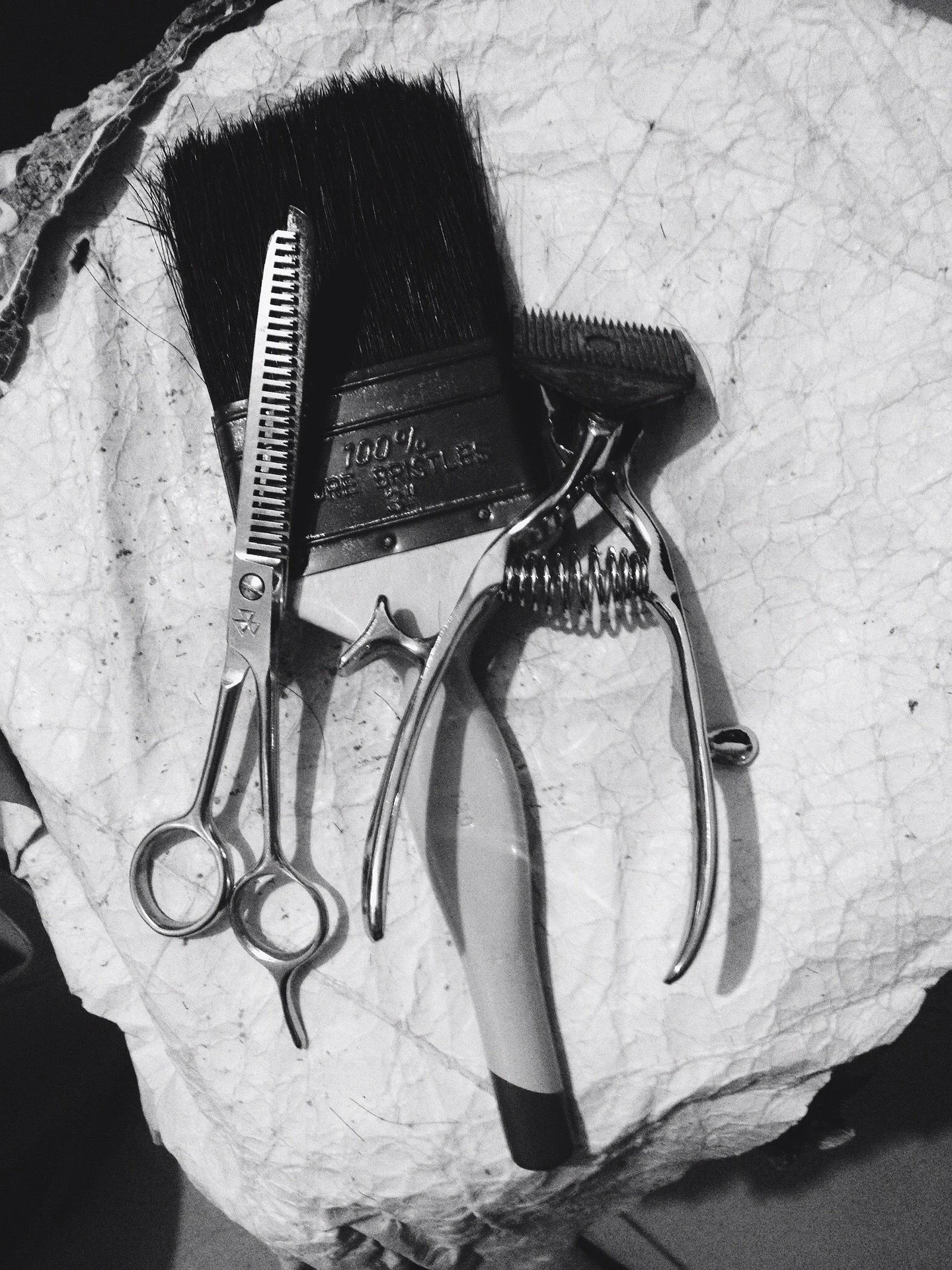 A pair of hair cutting scissors, a thinning scissors, a hair clipper, and a hairbrush are placed on a crumpled paper surface.
