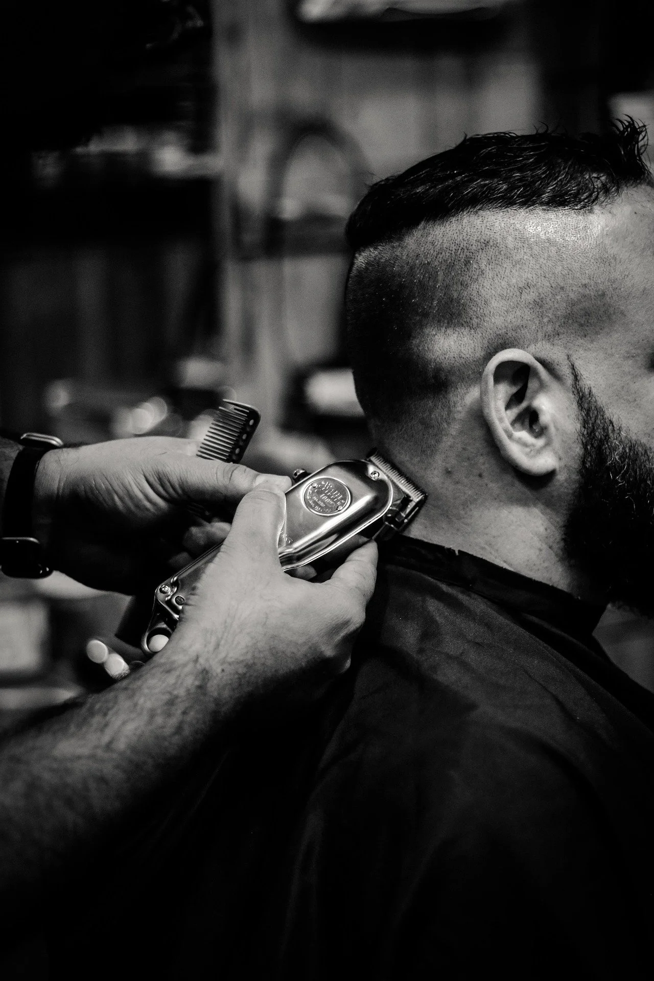 A man with a short haircut and beard getting a haircut. A barber is using clippers to trim the man's hair in a barbershop.
