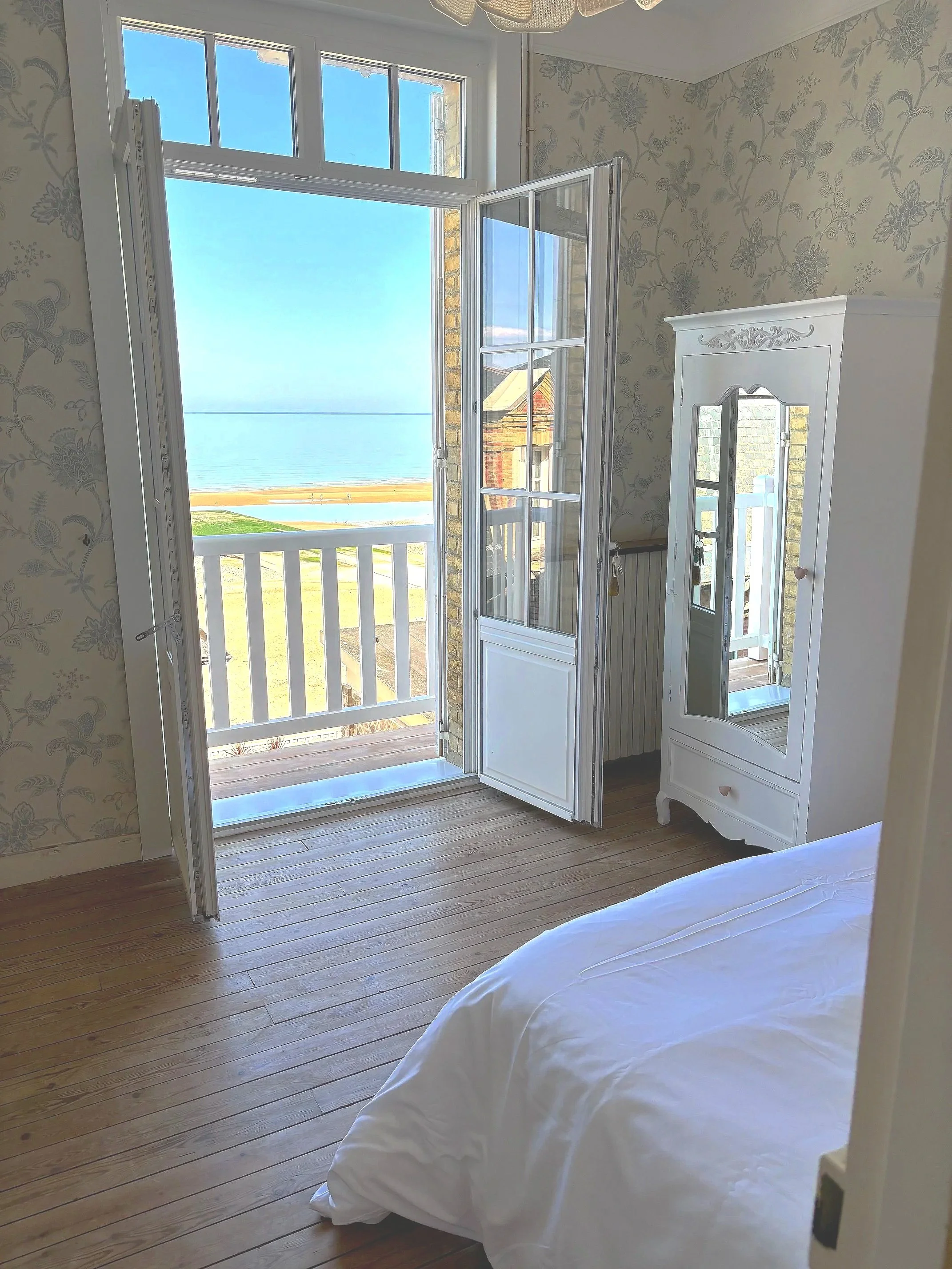 Chambre avec lit blanc et vue sur la mer à travers une grande fenêtre ouverte, mobilier blanc et murs avec papier peint floral.