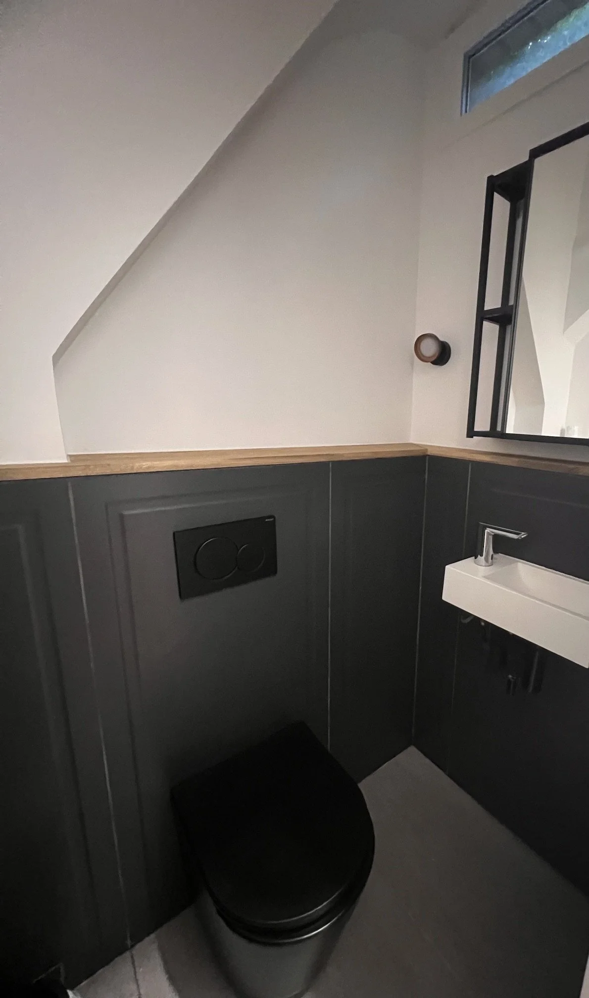 Petite salle de bain moderne avec un toilette noir, un lavabo blanc, un miroir, un mur en panneaux gris foncé, et un éclairage mural. Il y a aussi une fenêtre au plafond.