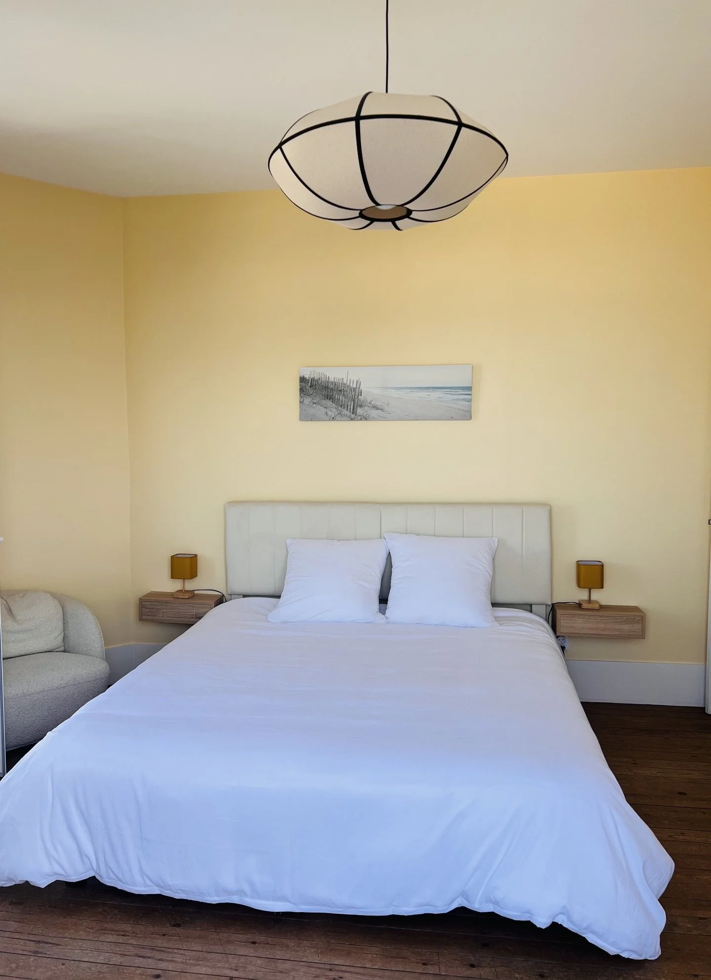 Chambre à coucher avec un lit blanc, deux tables de nuit avec lampes jaunes, un tableau représentant une plage, et un fauteuil gris à gauche.