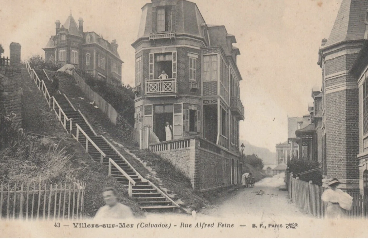Photographie ancienne montrant une rue pavée à Villers-sur-Mer, Calvados, avec des maisons en pierre et à tourelles. Des personnes sont visibles, certaines dans la rue, d'autres sur des escaliers montants sur la colline.
