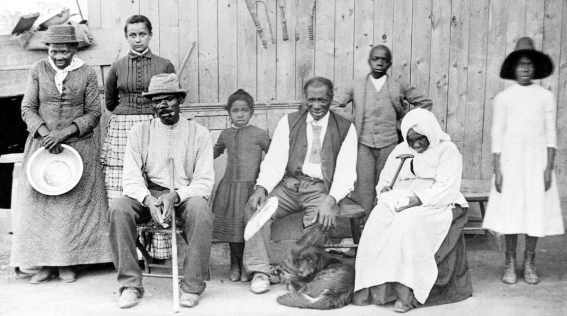 Historical black and white photograph of seven African American people and a dog outdoors against a wooden fence, including children, men, and women, some sitting and some standing.