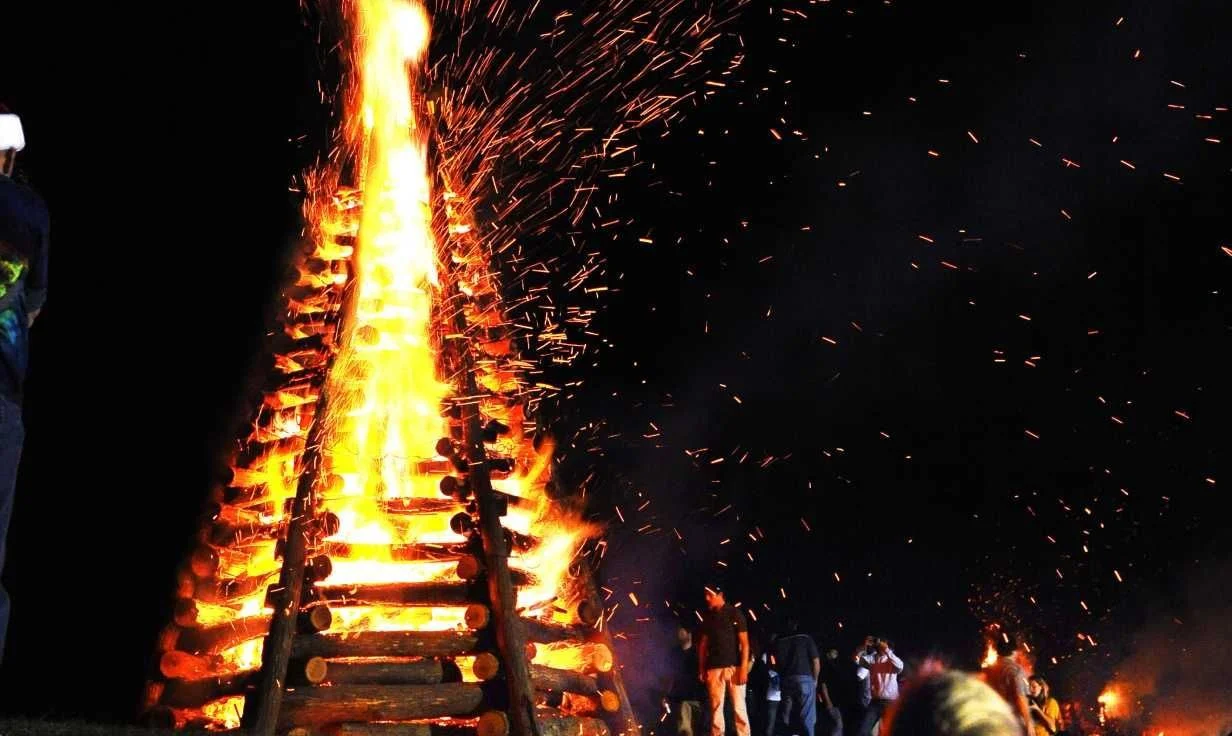 A traditional Louisiana Christmas Eve bonfire burns along the Mississippi River levee, illuminating the night sky with towering flames and sparks as spectators gather to celebrate a cherished holiday tradition.