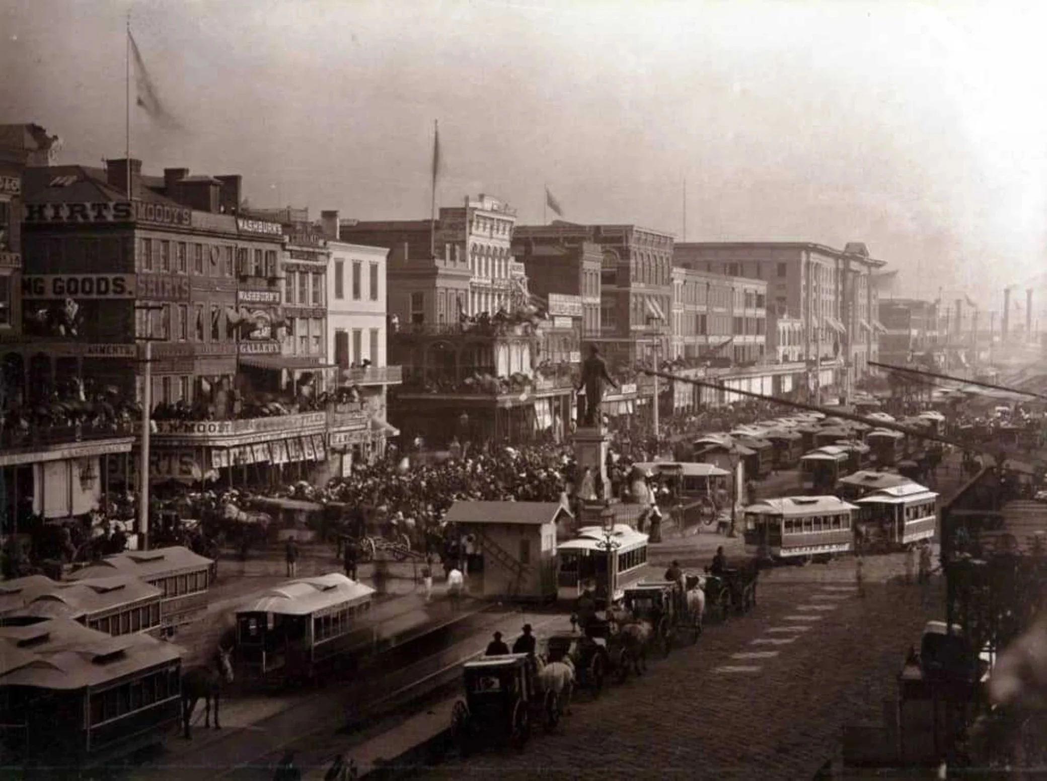 Historical black and white photograph of a busy downtown street filled with vintage streetcars, cars, and pedestrians, featuring multi-story commercial buildings with signs and awnings from the early 20th century.