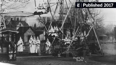 A photograph of an old amusement park or fairground ride with a large Ferris wheel and several small house-like structures in the background. The image appears to be black and white, and a group of people dressed in period clothing stand near the ride.