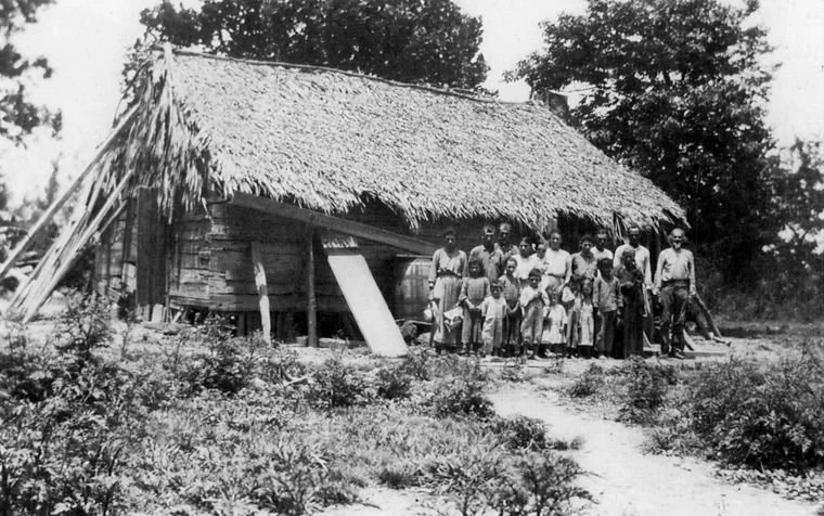A black-and-white photo of a group of people, including children, standing in front of a thatched-roof wooden house in a rural setting with trees and bushes.