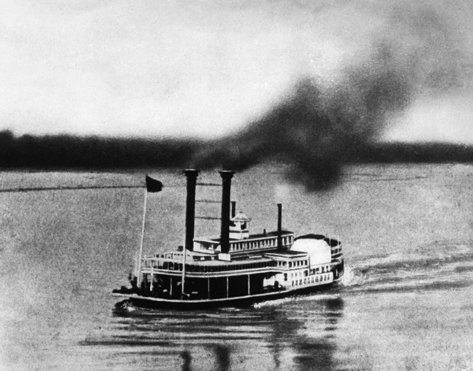 A black-and-white photo of a steamboat with three smokestacks on a body of water, emitting dark smoke, with a flat landscape in the background.