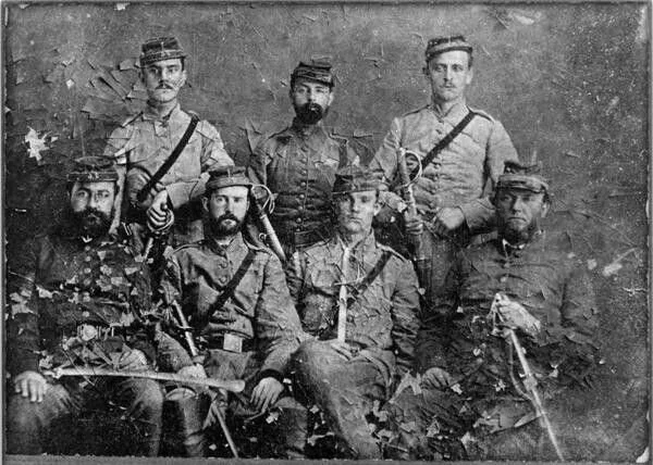 Group of seven men dressed in Civil War-era military uniforms, some holding swords or pistols, posed for a photo in front of a painted backdrop.