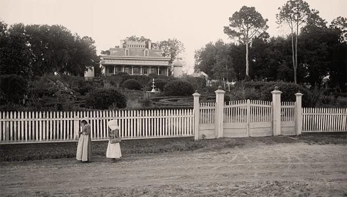 Black and white photo of two young girls standing near a white picket fence in front of a large historic house surrounded by trees, with a garden and fountain in the background.
