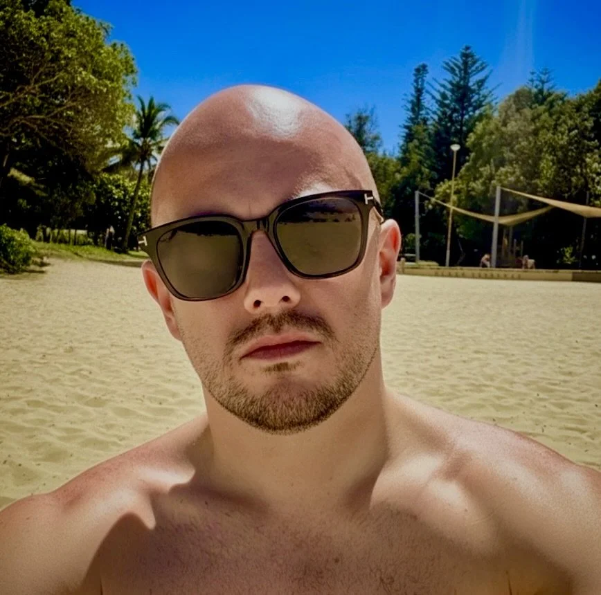 A man with a bald head, sunglasses, and a goatee taking a selfie at the beach with sand, trees, and a volleyball net in the background.