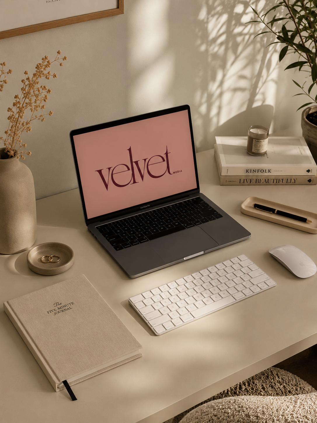 A modern workspace with a MacBook displaying 'Velvet Media' on the screen, a white keyboard, a mouse, a tray with a pen, a candle, a stack of books, a vase with dried flowers, and potted plant, and a beige journal on a white desk.