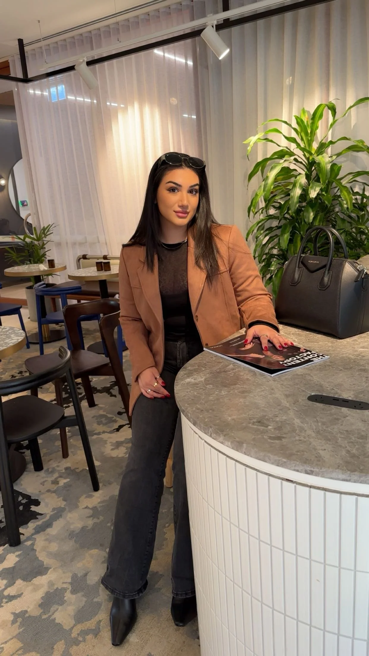 A woman with long dark hair, wearing a tan blazer, black top, and black jeans, sitting at a round marble table in a modern cafe with green plants and various colorful chairs in the background. She has sunglasses on her head and is holding a magazine.