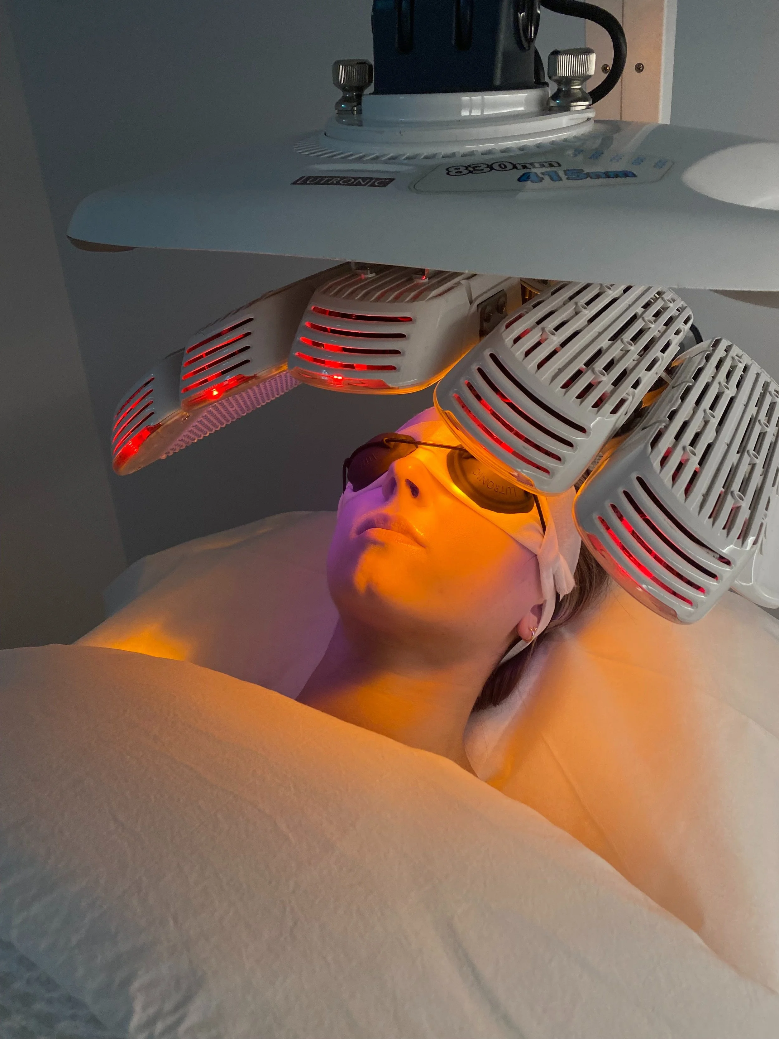 Person undergoing a red light therapy treatment with a large medical device covering the face, emitting orange and red light.