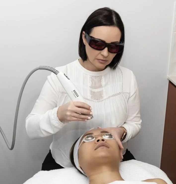 A woman wearing protective glasses performs a facial treatment with a handheld device on a woman's face in a clinical setting.