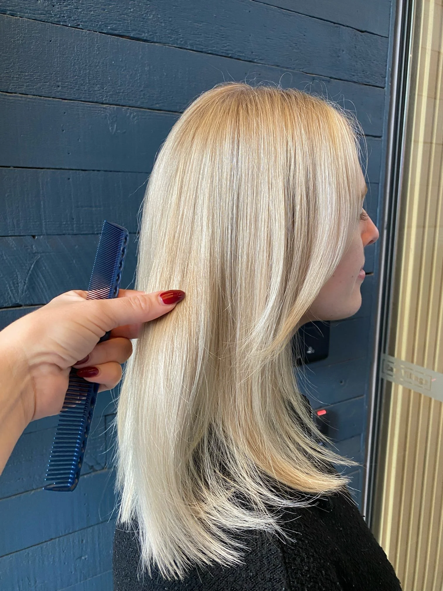 A woman with long, straight, platinum blonde hair in a salon, with a hairstylist holding a blue comb nearby.