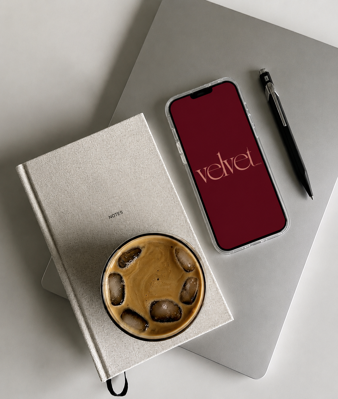 A flat lay of a workspace with a silver laptop, a black pen, a smartphone with a red wallpaper displaying the word 'velvet', a white notebook labeled 'NOTES', and a glass of iced coffee on a white table.