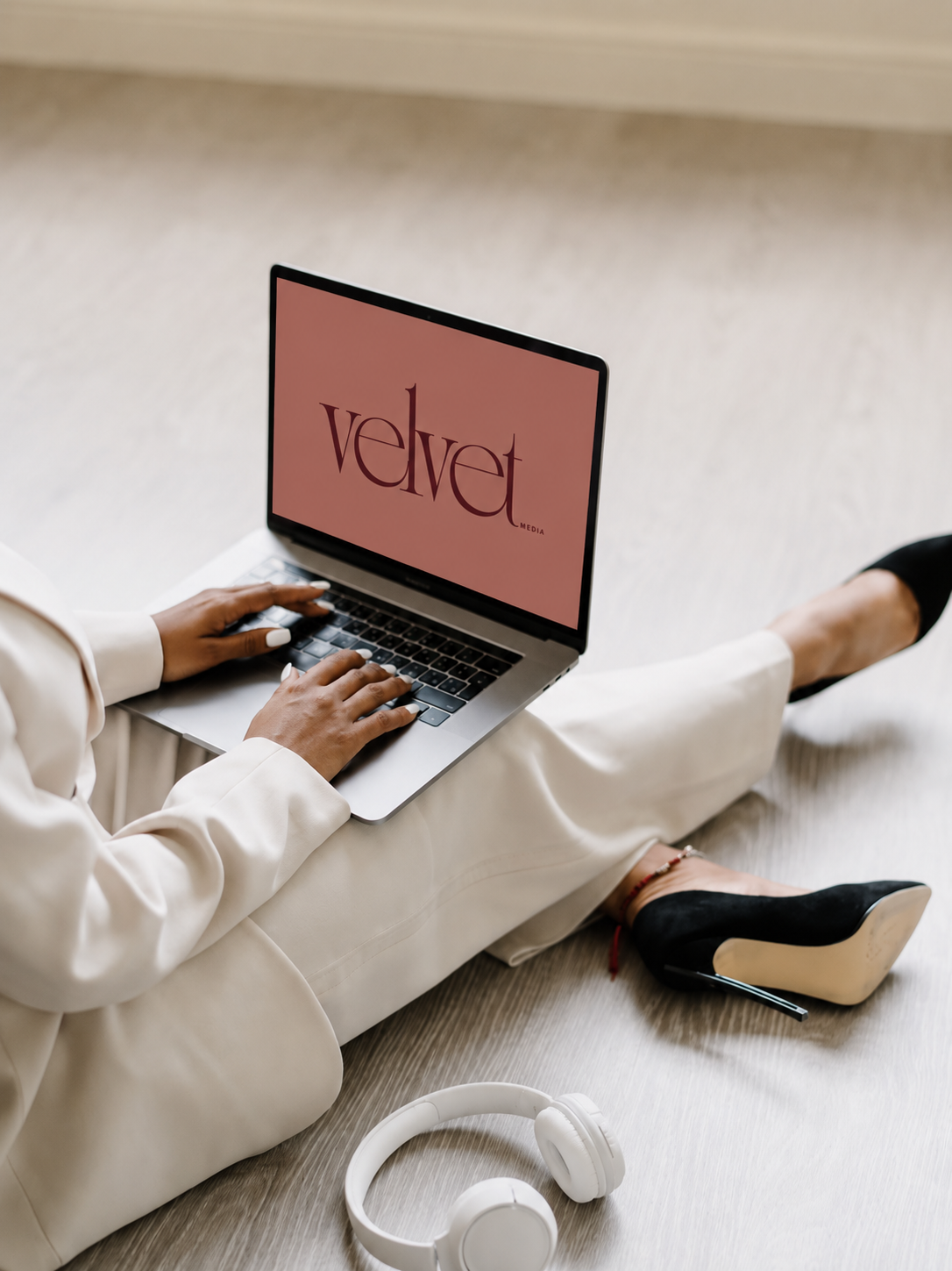 Person sitting on the floor using a laptop with a pink screen displaying "velvet media" logo, wearing cream blazer and pants, black high heels, with white headphones on the floor nearby.